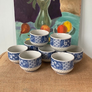 May include: A set of eight ceramic tea cups. Each cup features a white interior and a blue and white floral and geometric pattern around the exterior. The cups are stacked on a burlap surface.