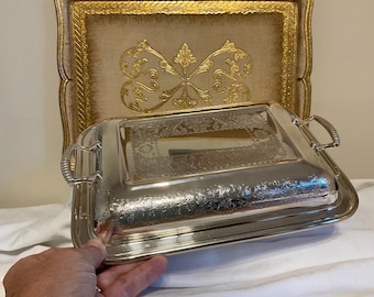 Può includere: Un piatto da portata in argento con un design decorato e un coperchio. Il piatto ha un bordo rialzato e due manici sui lati. Il coperchio ha un design simile al piatto e si adatta perfettamente sulla parte superiore.
