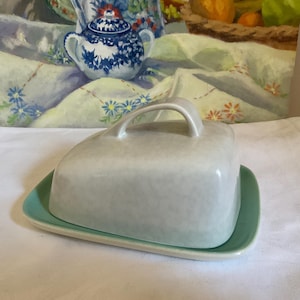 Op de afbeelding: Een lichtgrijze keramische botervloot met een handvat aan de bovenkant, rustend op een rechthoekige, lichtgroene basis. De schaal heeft een gespikkelde textuur. De achtergrond toont een schilderij met een blauw-witte theepot.