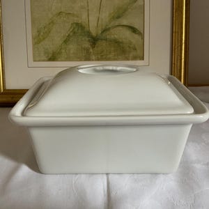May include: A white ceramic rectangular covered dish. The dish has a square shape with rounded corners and a matching lid with a small handle. The dish is sitting on a white tablecloth, with a framed picture in the background.
