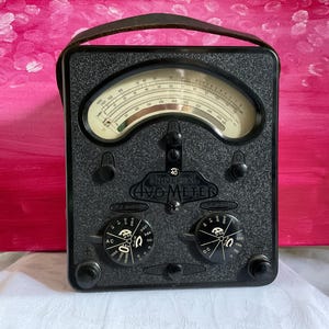 May include: A vintage black Universal AVO Meter with a leather handle. The meter features a curved gauge with a white face and black markings. Two dials are below the gauge, labelled AC and DC. This is a classic piece of electronic equipment.