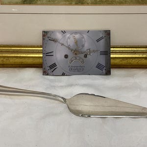 May include: A silver cake server with a decorative handle and a wide, flat blade. A vintage clock face with Roman numerals and ornate details is behind the server. The clock face has a floral border and the text "K. Summerhays, Canterbury".