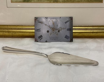May include: A silver cake server with a decorative handle and a wide, flat blade. A vintage clock face with Roman numerals and ornate details is behind the server. The clock face has a floral border and the text "K. Summerhays, Canterbury".