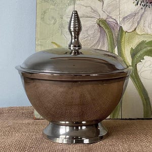 Può includere: Una zuppiera d'argento con coperchio e un pomo decorativo in cima. La zuppiera è rotonda e ha una base larga. La zuppiera è appoggiata su una superficie di juta marrone.