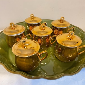 Puede incluir: Un juego de seis tazas de té verde oliva con tapas y asas doradas, dispuestas sobre una bandeja de servicio verde a juego con bordes festoneados. Las tazas y las tapas tienen un acabado brillante, lo que sugiere un estilo vintage.