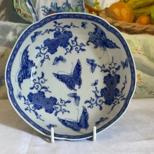 May include: A round ceramic bowl with a scalloped edge featuring a white base and a blue pattern of various butterflies and floral clusters. The rim is decorated with a geometric border, and the interior displays multiple butterfly illustrations.