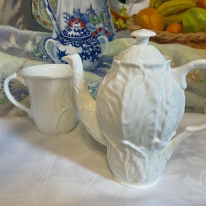 May include: A white ceramic teapot and matching mug featuring a raised, embossed cabbage leaf texture. The teapot has a lid with a small knob handle, a curved spout, and a handle. The mug has a matching leaf pattern and a curved handle.