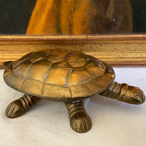 Puede incluir: Una figurita de tortuga de metal de color dorado con un caparazón y patas detalladas. La tortuga está sentada sobre una superficie blanca.