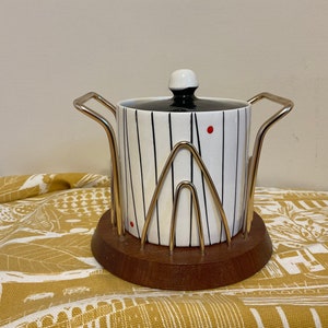 Può includere: Una zuccheriera in ceramica bianca con un design a strisce bianche e nere e punti rossi. La ciotola è su un supporto in metallo dorato con una base in legno.