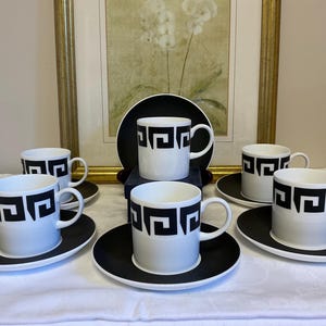 May include: A set of six white ceramic coffee cups and saucers. Each cup and saucer is black and white. The cups have a black geometric design around the top. The saucers are solid black. The set is arranged on a white tablecloth.