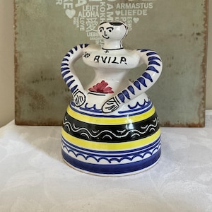 May include: A ceramic figurine of a woman with blue and white striped arms and a blue and white skirt with yellow and black accents. The figurine is holding a flower in her hand and has the word "AVILA" written on her chest.