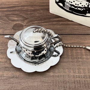 May include: Silver tea infuser shaped like a teapot with a chain and a flower-shaped base. The infuser has a lid and a strainer.