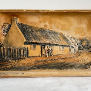 May include: A rectangular, tan-colored relief sculpture depicting a thatched-roof cottage with a fence and figures in front. The artwork is framed and features the text "Burns Cottage Alloway Ayr" at the bottom.