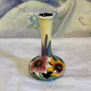 Può includere: Un piccolo vaso in ceramica con collo stretto e bordo svasato. La base presenta un motivo floreale dipinto a mano con grandi girasoli rosa e gialli, foglie verdi e spighe di grano viola su uno sfondo color crema e blu scuro.