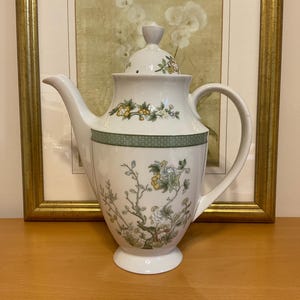 May include: White porcelain teapot with a lid, featuring a green band and floral design. The teapot has a curved spout and a large handle. The floral pattern includes yellow, green, and white flowers and branches. The teapot is on a wooden surface.