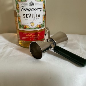 Peut inclure: Un jigger double en acier inoxydable avec un manche vert foncé est posé sur une surface blanche à côté d'une bouteille de gin Tanqueray Flor de Sevilla. La bouteille de gin a une étiquette rouge et une étiquette verte et jaune.