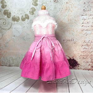 May include: A doll dress featuring a white lace bodice with a high collar and a pink tiered skirt. The skirt has an elastic waistband with a drawstring tie and a hemline decorated with embroidered spiral patterns in a matching pink thread.