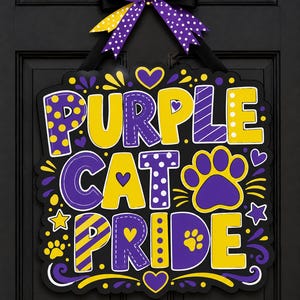May include: A black rectangular door sign featuring the text PURPLE CAT PRIDE in yellow and purple block letters with polka dot and striped patterns. The sign is decorated with purple paw prints, yellow stars, and hearts, topped with a purple and yellow bow.