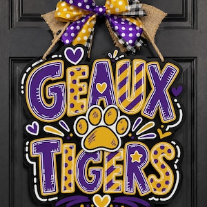 May include: A decorative wooden door sign featuring the text GEAUX TIGERS in purple and yellow with polka dot and striped patterns. A yellow paw print sits in the center, surrounded by small hearts. A multi-layered bow with burlap, polka dot, and plaid ribbons is attached to the top.