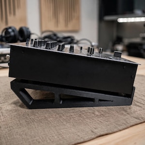 Può includere: Una console mixer audio nera poggia su un supporto nero angolato a telaio aperto. Il mixer presenta una superficie superiore piatta con numerosi manopole di controllo e cursori neri disposti in file. Il supporto ha un design geometrico cavo.