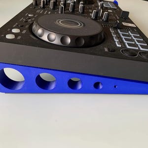 Può includere: Un controller DJ nero appoggiato su un supporto blu. Il controller ha varie manopole, pulsanti e una jog wheel centrale. Il supporto ha un design curvo con più fori circolari, che offrono supporto e un'estetica moderna.