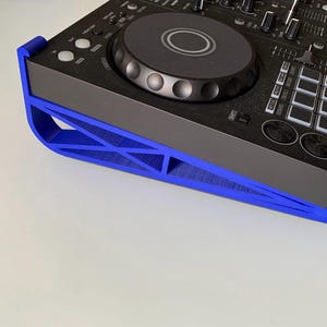 Puede incluir: Un controlador de DJ negro con un soporte azul. El controlador tiene una rueda central, botones y controles deslizantes. El soporte azul está en ángulo y tiene un diseño geométrico.