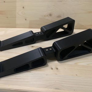 Könnte beinhalten: Zwei mattschwarze Laptop-Ständer mit geometrischem, offenem Rahmendesign und dreieckigen Aussparungen. Jeder Ständer hat eine hakenförmige Vorderkante zur Sicherung eines Geräts und besteht aus zwei verbundenen Segmenten, die mit zwei kleinen silbernen Metallschrauben befestigt sind.