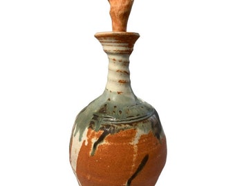 May include: A ceramic bottle with a rounded body and a tall, slender neck. The bottle is decorated with a mix of brown, white, and green glazes, creating a unique and abstract design. The bottle has a wooden stopper.