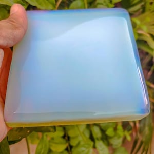 Könnte beinhalten: Ein quadratischer, irisierender, weißer Opalitkristall mit einer glatten, polierten Oberfläche. Der Kristall hat einen hellblauen, gelben und rosafarbenen Farbton.