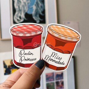 May include: Two red and orange glass jars with white lids and labels. The labels have black text that reads "Pectin Princess" and "Miss Marmalade".