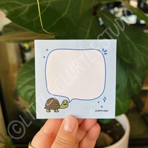 May include: A blue note card with a white speech bubble and a cartoon turtle. The speech bubble has a blue outline and the turtle has a brown shell and green head and feet. The text on the note card says "@creative.lagan".