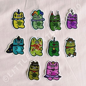 May include: A collection of colorful frog stickers with various designs. Each sticker features a cartoon frog in different outfits, including a wizard hat, a cowboy hat, and a party hat. The frogs are in shades of green, blue, yellow, and purple.