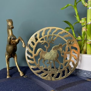 May include: A brass trivet with a circular design featuring a stylised horse and a tree. The trivet is a decorative piece with a cutout design.