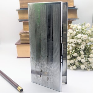 May include: A silver metal cigarette case with a textured surface and a hinged lid. The case is closed and standing upright on a table.