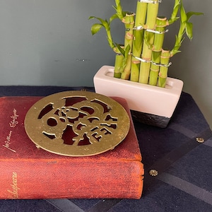 Puede incluir: Un posavasos de latón con un diseño decorativo de corte se encuentra sobre un libro encuadernado en cuero rojo. El posavasos es redondo y presenta un patrón floral con detalles intrincados. El libro está cerrado y tiene un título dorado en el lomo.