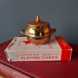 May include: A small gold pot with a copper lid sits on top of a pack of playing cards. The playing cards are red and white and say "Superior Quality Playing Cards".