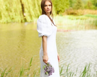 May include: A woman in a white linen dress with puffed sleeves stands by a pond holding a bouquet of purple flowers.