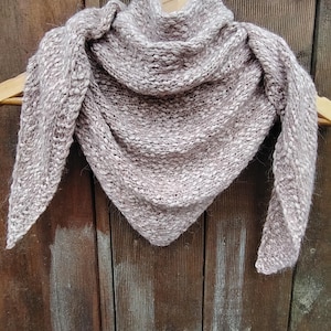May include: A soft, light pink knitted shawl with a triangular shape, hanging on a wooden hanger.