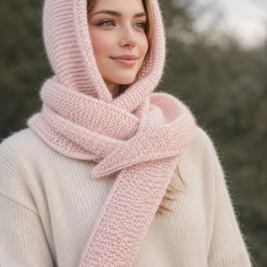 Könnte beinhalten: Ein hellrosa gestrickter Kapuzenschal mit strukturiertem Strickmuster. Das Accessoire besteht aus einer Kapuze, die in einen langen, wickelbaren Schal übergeht und durch das weiche, gleichmäßige Strickgewebe Kopf und Hals vollständig bedeckt.