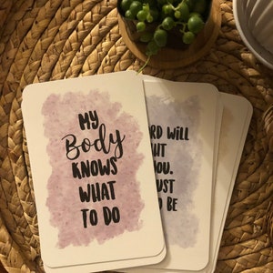 May include: A stack of white cards with rounded corners, each featuring inspirational text in black font against a watercolor-style background. The card on top reads "MY BODY KNOWS WHAT TO DO". The cards rest on a woven, circular placemat.