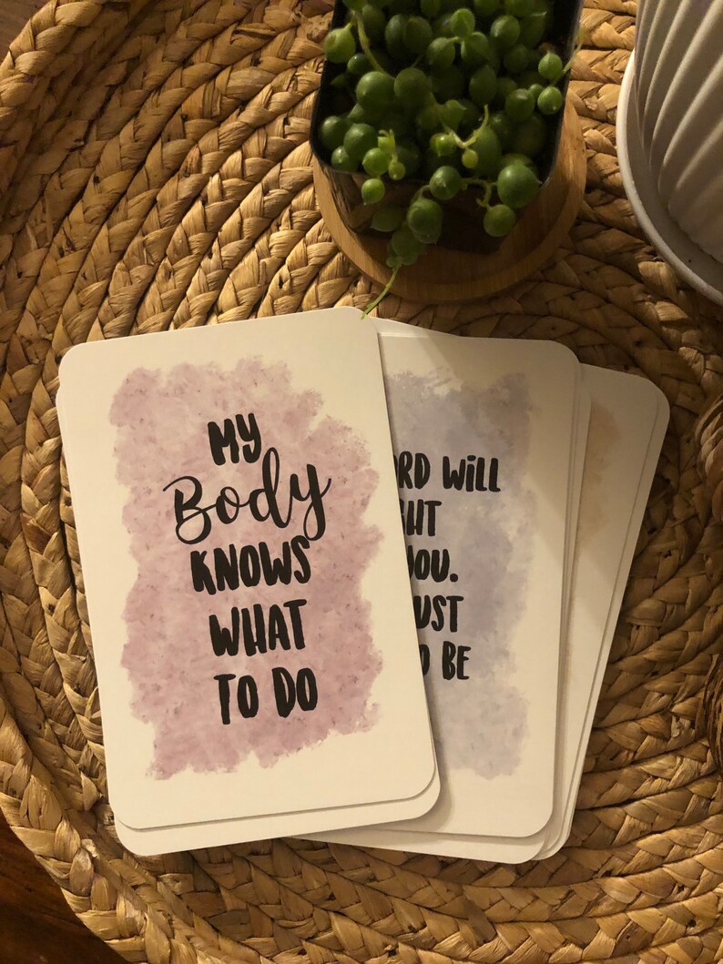 May include: A stack of white cards with rounded corners, each featuring inspirational text in black font against a watercolor-style background. The card on top reads "MY BODY KNOWS WHAT TO DO". The cards rest on a woven, circular placemat.