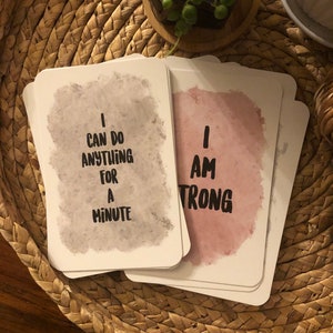 May include: A stack of white affirmation cards with rounded corners. One card reads "I CAN DO ANYTHING FOR A MINUTE" in black text over a gray watercolor wash. Another card reads "I AM STRONG" in black text over a pink watercolor wash.