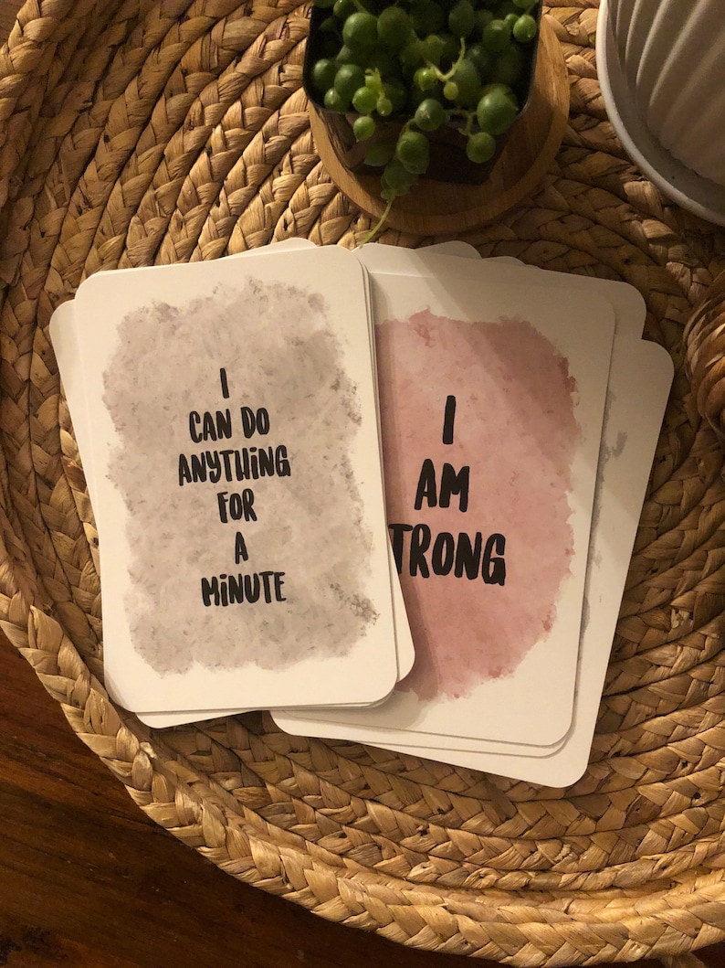 May include: A stack of white affirmation cards with rounded corners. One card reads "I CAN DO ANYTHING FOR A MINUTE" in black text over a gray watercolor wash. Another card reads "I AM STRONG" in black text over a pink watercolor wash.