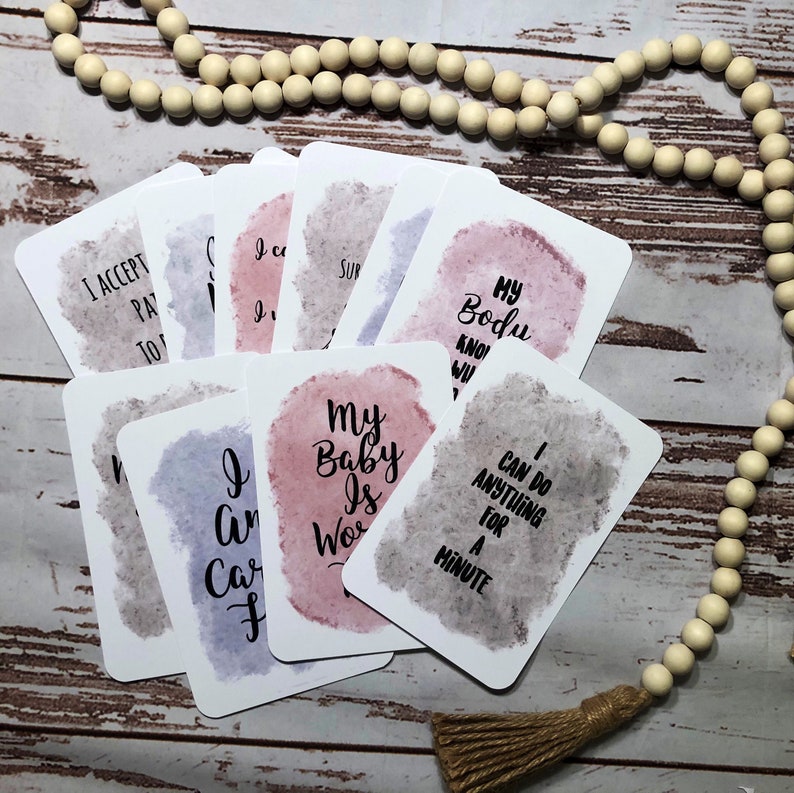 May include: A collection of affirmation cards with watercolor backgrounds in shades of pink, purple, and gray. Each card features a different positive affirmation in black script. A wooden bead garland and tassel are in the background.