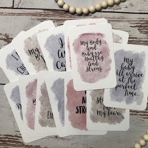 May include: A collection of affirmation cards with watercolor backgrounds in shades of purple, pink, and gray. Each card features inspirational text in a handwritten style font. The cards are arranged on a light wood surface.
