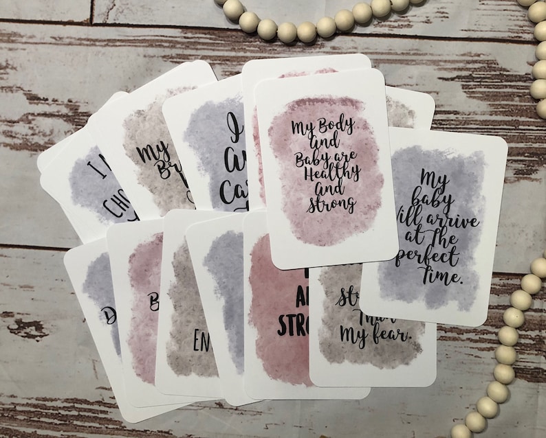 May include: A collection of affirmation cards with watercolor backgrounds in shades of purple, pink, and gray. Each card features inspirational text in a handwritten style font. The cards are arranged on a light wood surface.