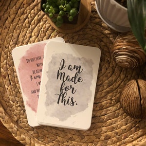May include: A stack of white affirmation cards with rounded corners. One card has the text "I am Made for This" in black script over a gray watercolor wash. Another card has text over a pink watercolor wash. The cards rest on a woven tray.