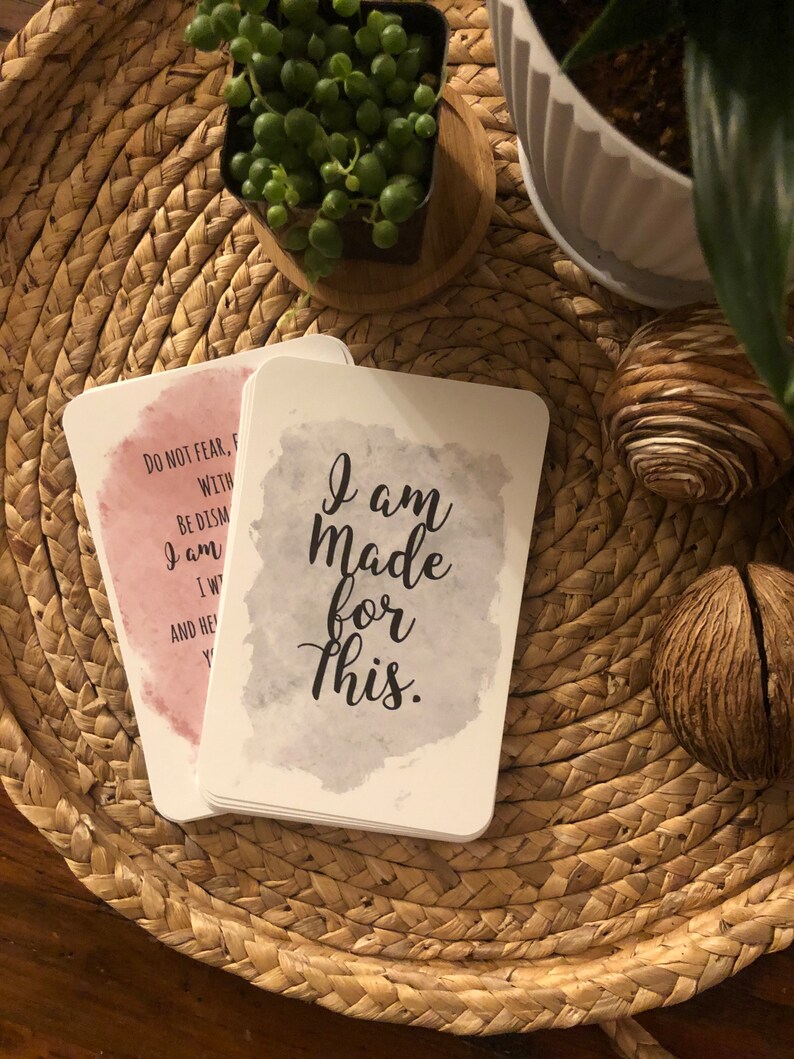 May include: A stack of white affirmation cards with rounded corners. One card has the text "I am Made for This" in black script over a gray watercolor wash. Another card has text over a pink watercolor wash. The cards rest on a woven tray.