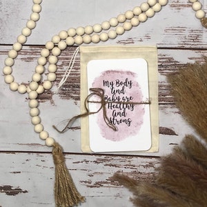 May include: A beige canvas pouch with a card that reads "My Body and Baby are Healthy and Strong" tied with twine. A wooden bead garland and dried pampas grass are also in the image.