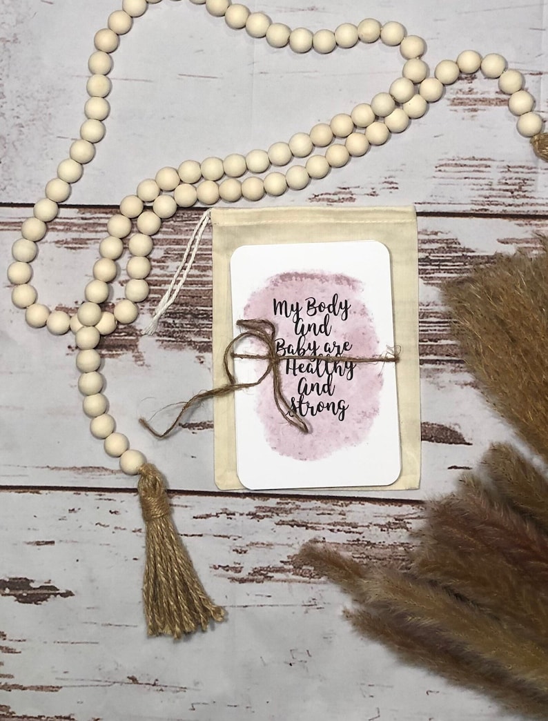 May include: A beige canvas pouch with a card that reads "My Body and Baby are Healthy and Strong" tied with twine. A wooden bead garland and dried pampas grass are also in the image.